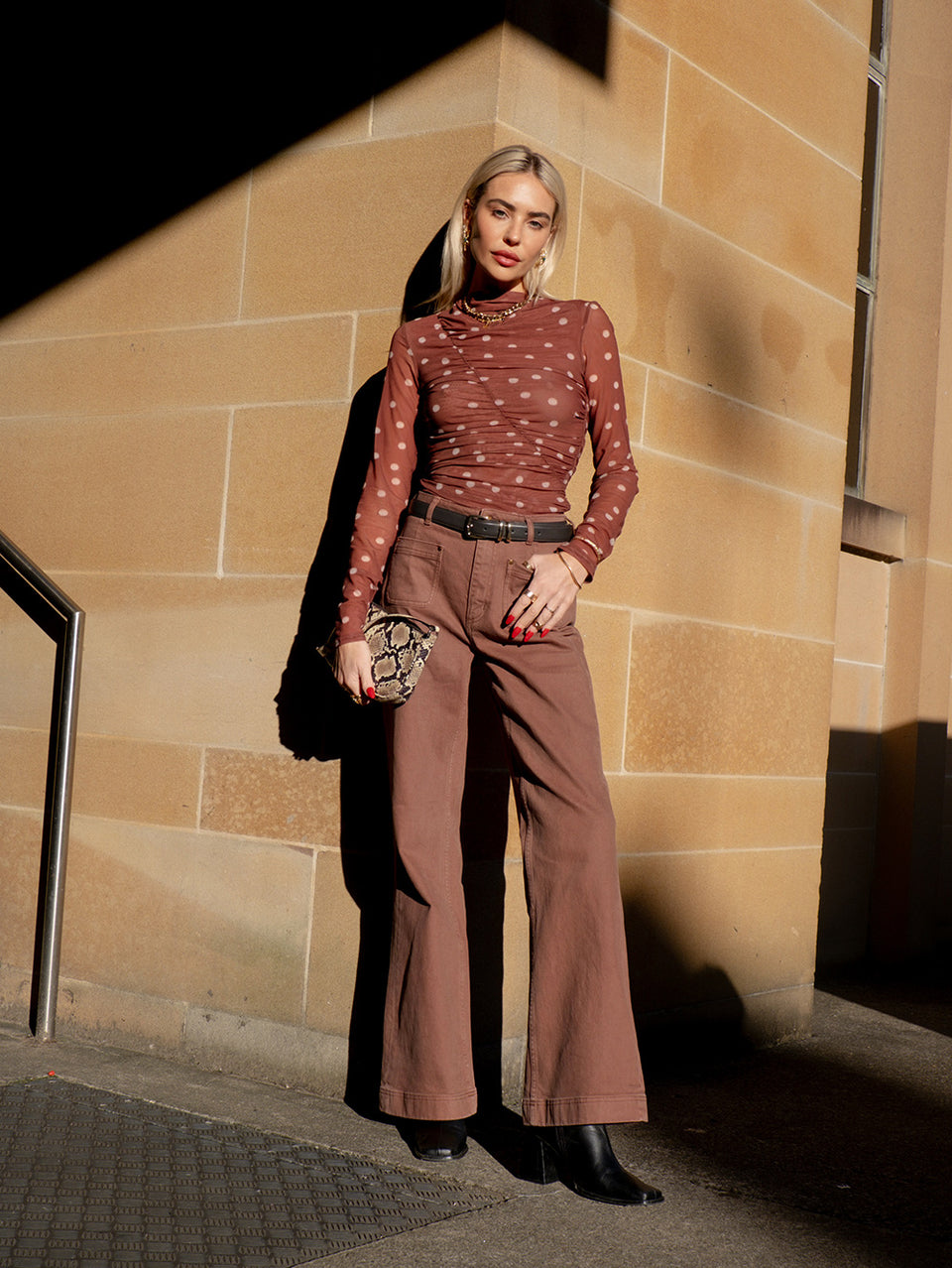 KIVARI influencer lifestyle shot - model wearing brown polka dot long-sleeve mesh top with brown wide-leg trousers and snake-print bag