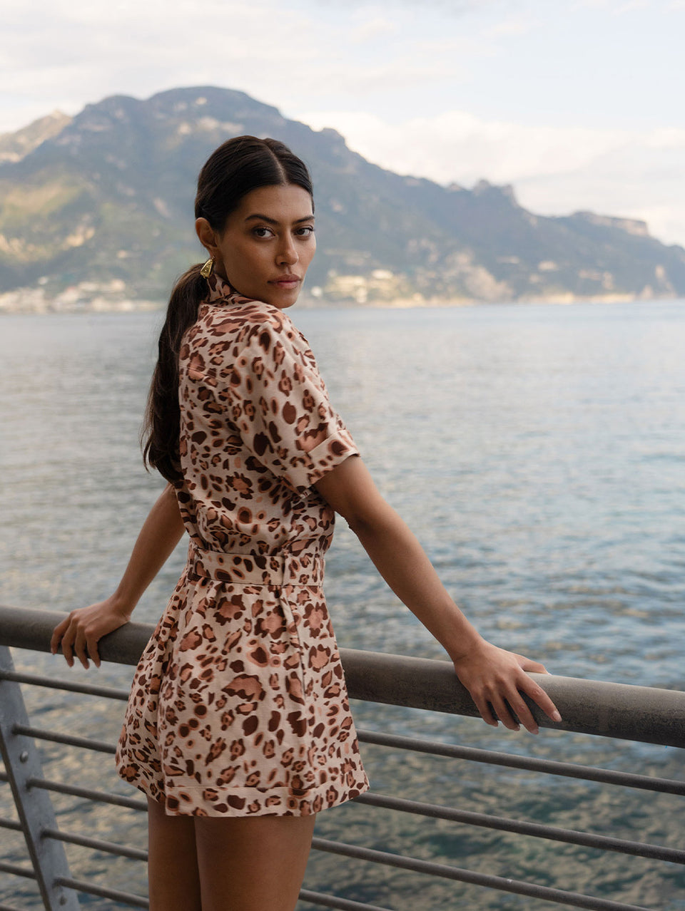 Woman in a leopard print playsuit standing by a railing with mountains and water in the background