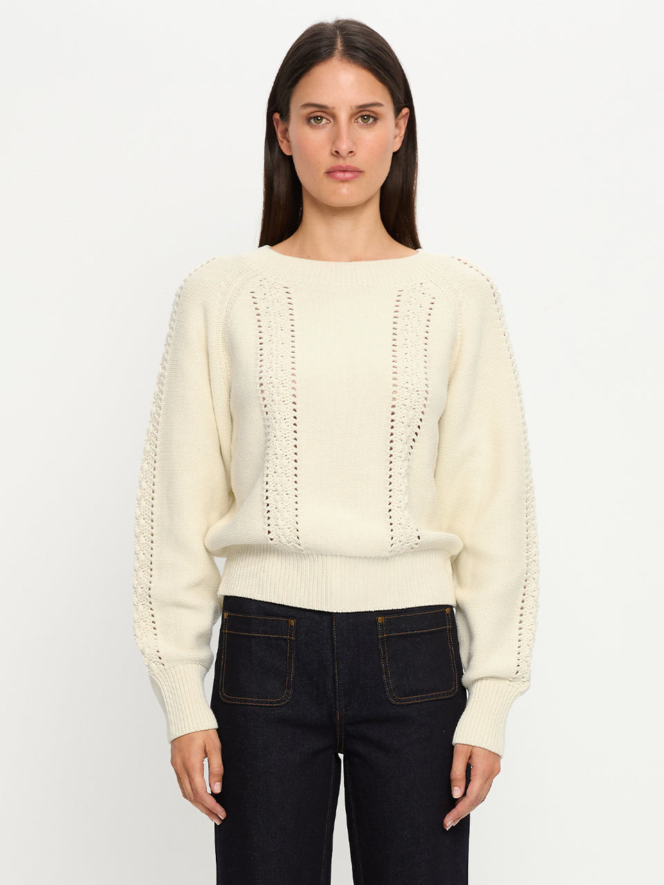 Woman wearing a cream sweater and dark jeans against a white background
