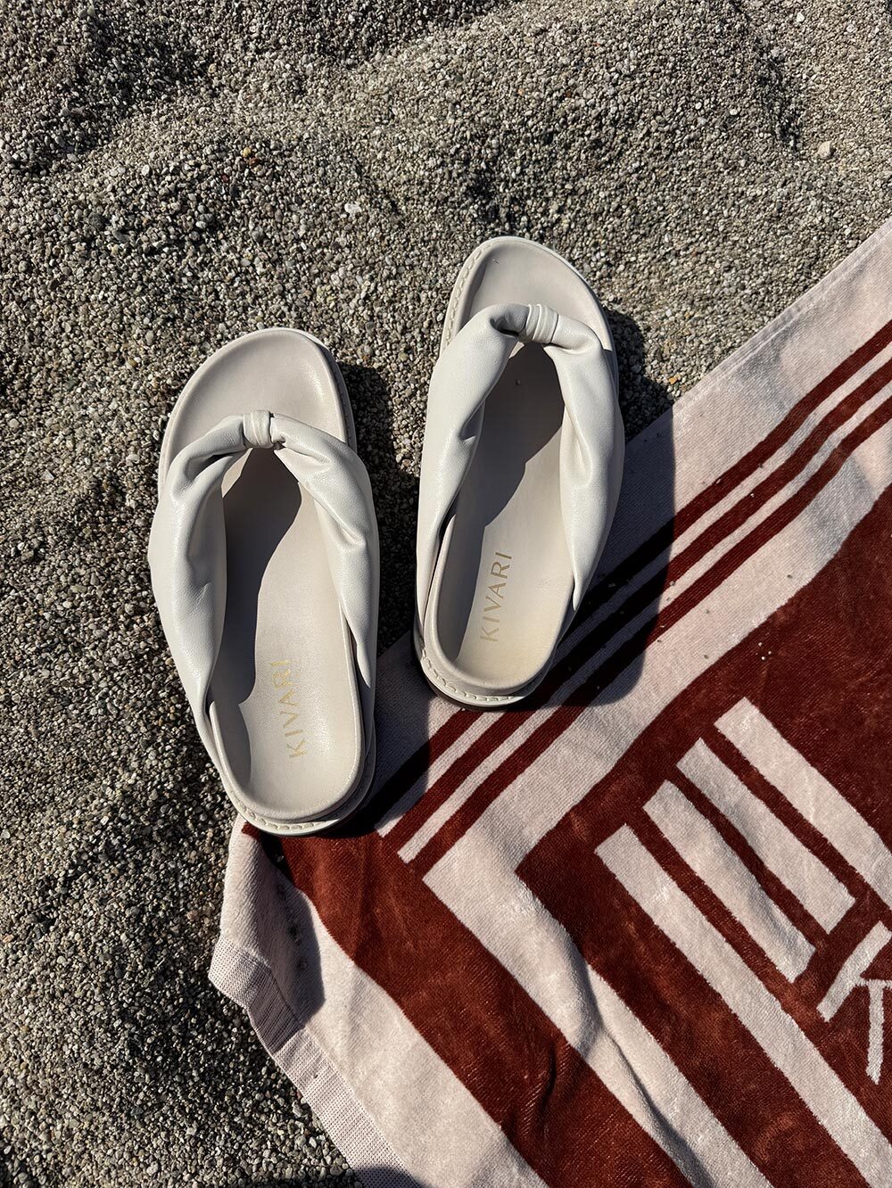 Pair of light creamsandals on a towel with a KIVARI logo on a sandy beach