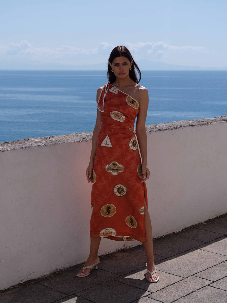 Woman in an orange dress with geometric patterns standing on a rooftop with ocean view